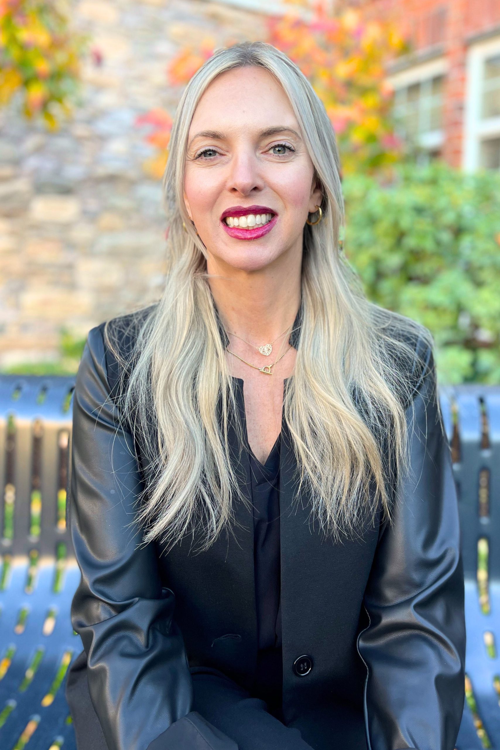 Lisa Cianni sitting outside in front Laurel Parc Senior Living Community Wearing a Black Leather Jacket in front of autumn leaves